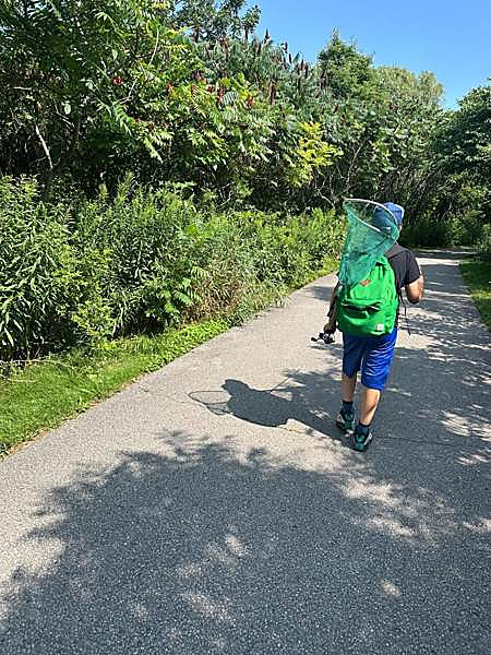 A child walking a city trail with a net over one shoulder and a fishing rod in hand