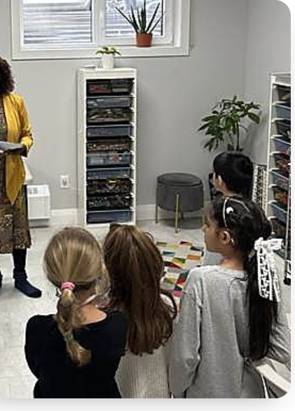 A guide in a yellow cardigan talking with three girls gathered around a colourful mat, craft supplies on the shelves behind them