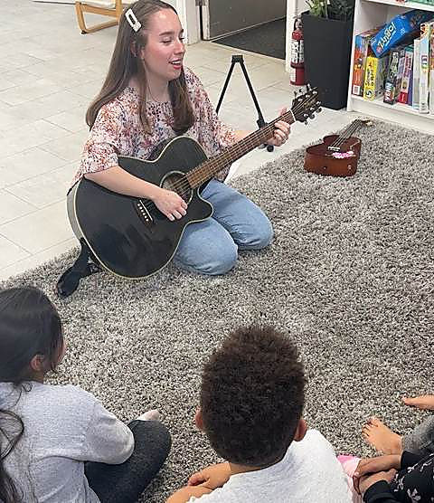 A Hexagon guide playing guitar to a group of kids sitting on the rug
